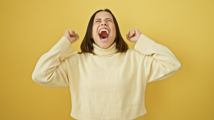Energetic young hispanic woman cheering in a cream sweater against a bright yellow background.