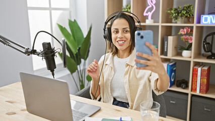 A smiling young woman with headphones takes a selfie in a modern radio studio setup, showcasing...