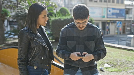 A man using smartphone by a woman, both standing by a city street with buildings and a road in the...