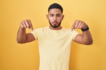 Young hispanic man standing over yellow background pointing down looking sad and upset, indicating...