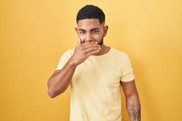 Young hispanic man standing over yellow background laughing and embarrassed giggle covering mouth...