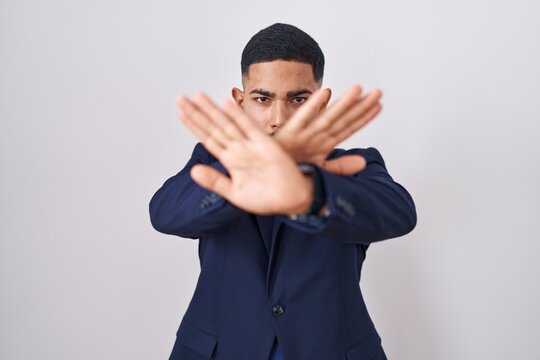 Young hispanic man wearing business suit and tie rejection expression crossing arms and palms doing negative sign, angry face