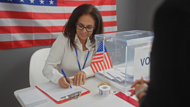 A woman volunteering at a united states electoral college writes notes under an american flag indoors.