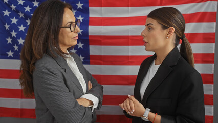 Two professional women in conversation against a backdrop of an american flag in an office interior.