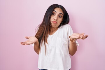Young arab woman standing over pink background clueless and confused expression with arms and hands...