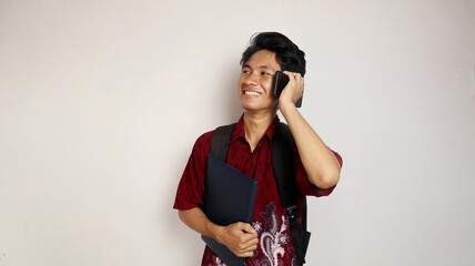 happy young handsome asian man in batik shirt holding a bag and carrying a book posing while making...