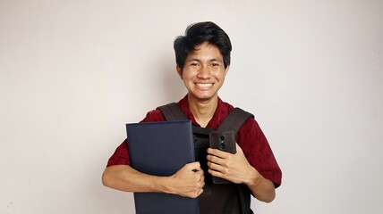 Handsome young Asian man in batik shirt carrying a bag, holding a book and smartphone, expressing...