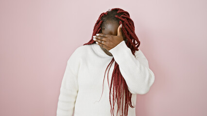 African american woman with braids wearing a sweater covers her face over a pink background