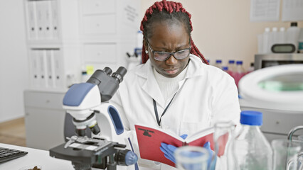 A focused black woman with braids reads a book in a laboratory, showcasing education and...
