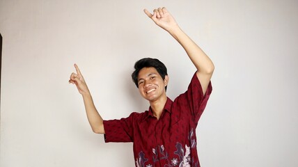 Excited handsome Asian young man wearing a batik shirt and smiling friendly pointing upwards with...