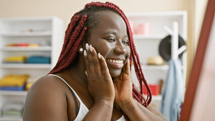 A happy african woman with braids touches her face while looking in the mirror in a clothing room.
