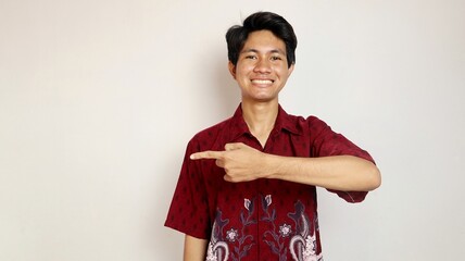 Excited handsome Asian young man wearing a batik shirt and smiling friendly pointing to the side