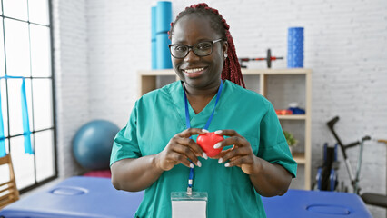 African american woman with braids smiling in scrubs holding a heart inside a bright physiotherapy...