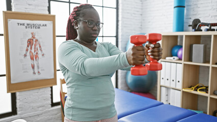 African american woman with braids lifting dumbbells in a physiotherapy clinic interior.
