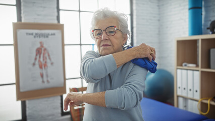 A senior woman applies ice to her shoulder in a physiotherapy clinic with anatomical posters,...