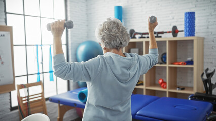 Senior woman exercises with dumbbells in a bright physiotherapy clinic showing fitness, health, and...