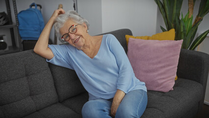 A smiling elderly woman with grey hair wearing glasses relaxes on a couch in a cozy living room.