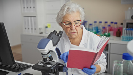 Senior woman scientist reading a book while working with a microscope in a laboratory setting.