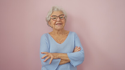 A smiling senior woman with grey hair and glasses stands arms crossed in front of a pink wall...