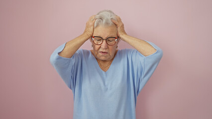 Senior woman with glasses holding head in frustration against a plain pink background.