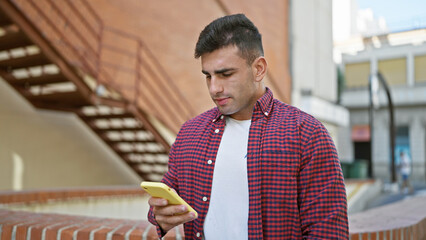Cool hispanic guy, portrait of a serious young man engrossed in texting on his smartphone, standing...