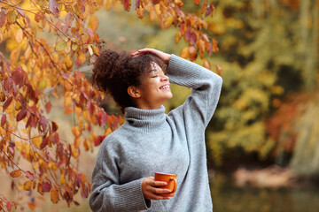 Young happy smiling mixed-race woman with coffee cup in autumn nature, pleased african american female with curly hair in knitted sweater