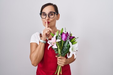 Middle age brunette woman wearing apron working at florist shop holding bouquet asking to be quiet...