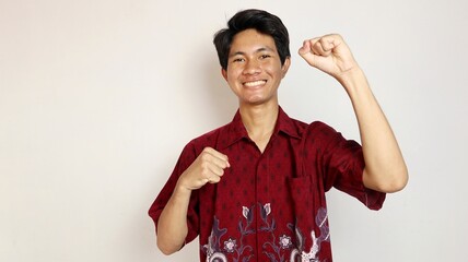 Handsome Asian young man dressed in batik posing with both hands raised, very excited and happy