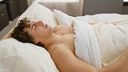 A young hispanic man with curly hair is peacefully sleeping shirtless in a comfortable home bedroom setting