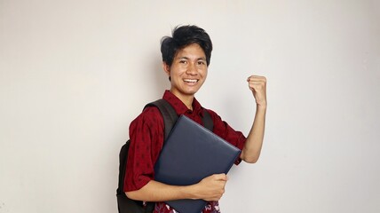 Handsome Asian young man dressed in batik holding bag holding books posing excitedly