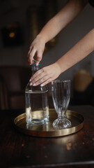 Close-up of a woman's hands unscrewing a bottle of water on a tray with an empty glass in a dark cozy restaurant setting.