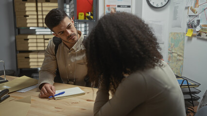 A man interviews a woman in an office filled with investigation evidence like photos, maps, and...