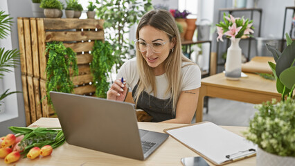 A focused blonde woman works on a laptop in a flower-filled indoor space, evoking a fresh,...