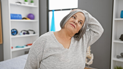 A senior woman stretches her neck in a physical therapy clinic, indicating healthcare, wellness,...
