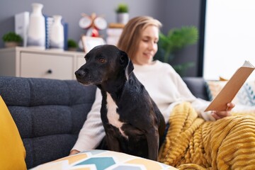 Young blonde woman reading book sitting on sofa with dog at home