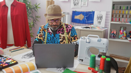 A stylish tailor in a hat reviews designs on a laptop in his colorful atelier, surrounded by sewing...