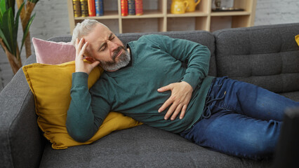A middle-aged bearded man lying on a couch at home, appearing to suffer from stomach pain.