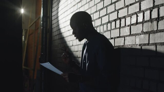 Lowkey medium shot of young Black male standup comedian rehearsing his monologue and laughing while standing in dark against brick wall corridor before going on stage