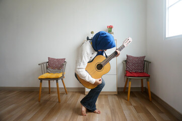 Full length portrait of excited Asian muslim female musician in hijab playing an acoustic guitar in...