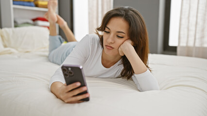 Hispanic woman lying on a white bed using a smartphone in a bright bedroom.