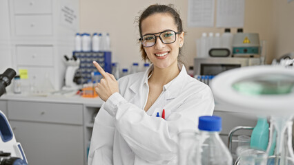 A smiling woman scientist pointing at laboratory equipment indoors.
