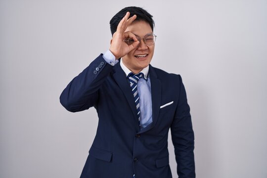 Young asian man wearing business suit and tie doing ok gesture with hand smiling, eye looking through fingers with happy face.