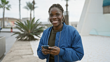 A smiling african american woman with braids using a smartphone on a city street.