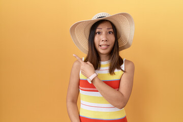 Middle age chinese woman wearing summer hat over yellow background pointing aside worried and...