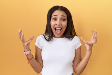 Young arab woman wearing casual white t shirt over yellow background crazy and mad shouting and...