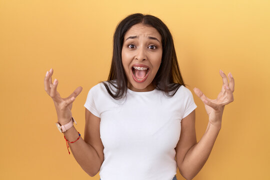 Young arab woman wearing casual white t shirt over yellow background crazy and mad shouting and yelling with aggressive expression and arms raised. frustration concept.