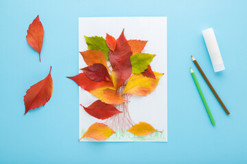 Tree shape created from colorful leaves on white paper, glue stick and pencils on light blue table background. Pastel color. Making autumn decorations. Closeup. Top view.
