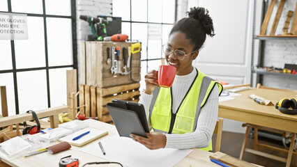 African american woman in safety vest sipping coffee and using tablet in carpentry workshop.