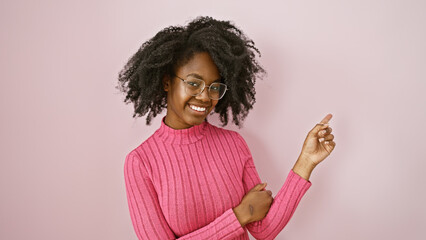 Smiling african woman in pink sweater points happily against a plain indoor background, exuding...