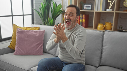 Bearded senior man clapping and smiling cheerfully in a cozy living room environment.